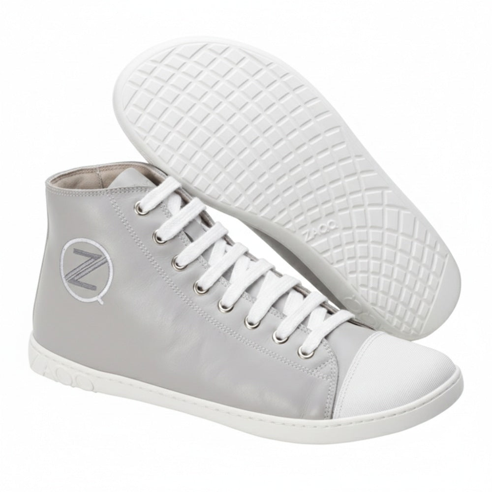 Ausgestellt sind die CHUQQS Grey High-Top-Sneaker von ZAQQ aus Nappaleder. Ein Schuh liegt auf der Seite und zeigt ein strukturiertes Sohlenmuster, das an einen Barfußschuh erinnert, während der andere aufrecht steht und Schnürsenkel und ein rundes Logo mit dem Buchstaben Z zeigt.