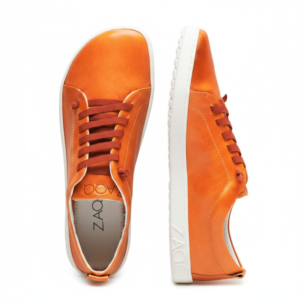 Wir präsentieren den STIQ Sierra von ZAQQ: Ein auffälliges Paar leuchtend orangefarbener Sneaker aus Nappaleder mit auffälligen braunen Schnürsenkeln und grauen Sohlen. Der linke Sneaker wird elegant von der Seite präsentiert, während der rechte von oben betrachtet wird und das markante Zaoo-Logo auf der Innensohle und entlang der Sohlenkante zeigt. Diese schicken Barfußschuhe vereinen mühelos Komfort und Stil.